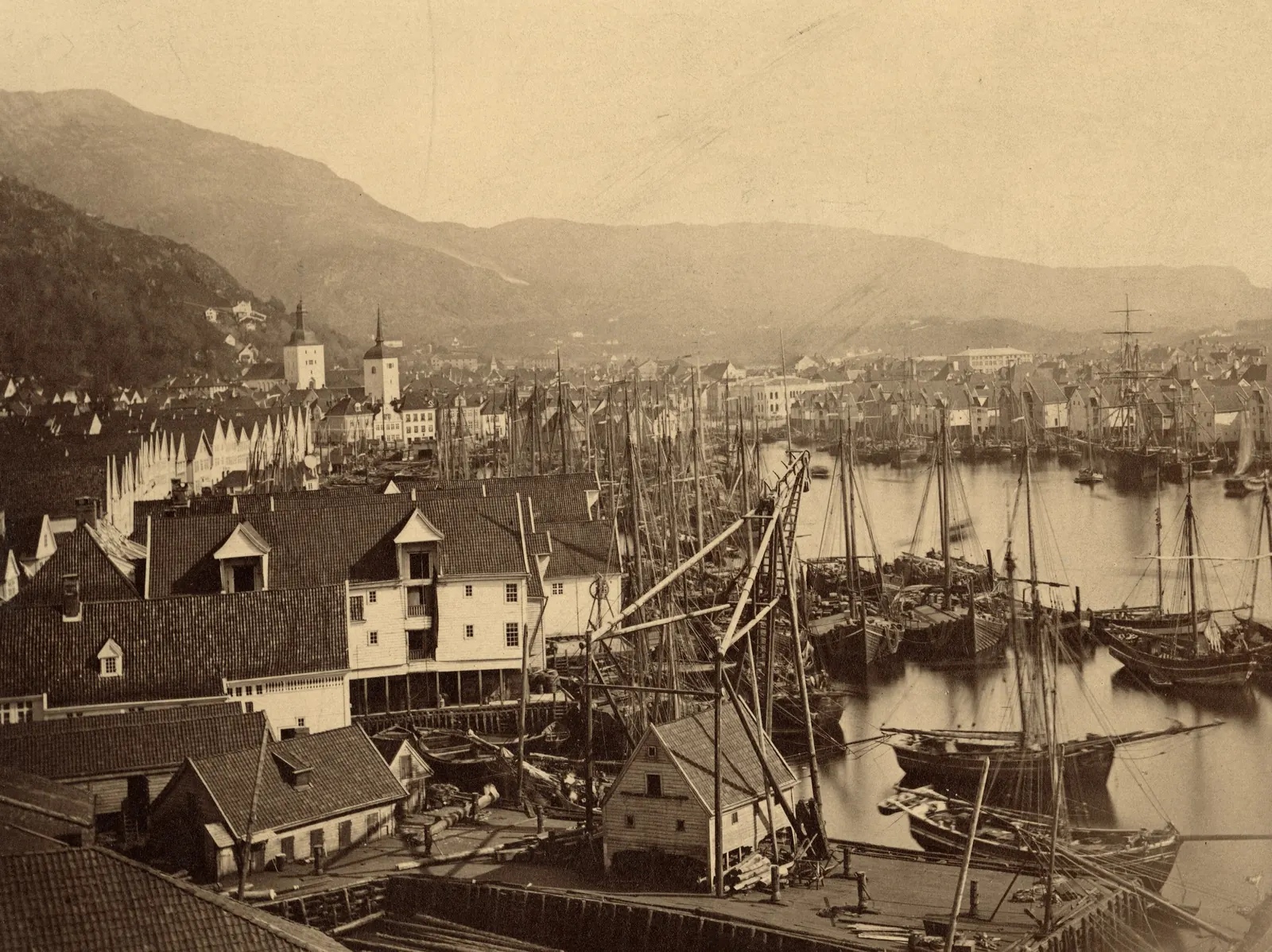 Nineteenth-century photograph of a harbor crowded with masts, a row of gabled wooden warehouses along the far quay, and mountains beyond