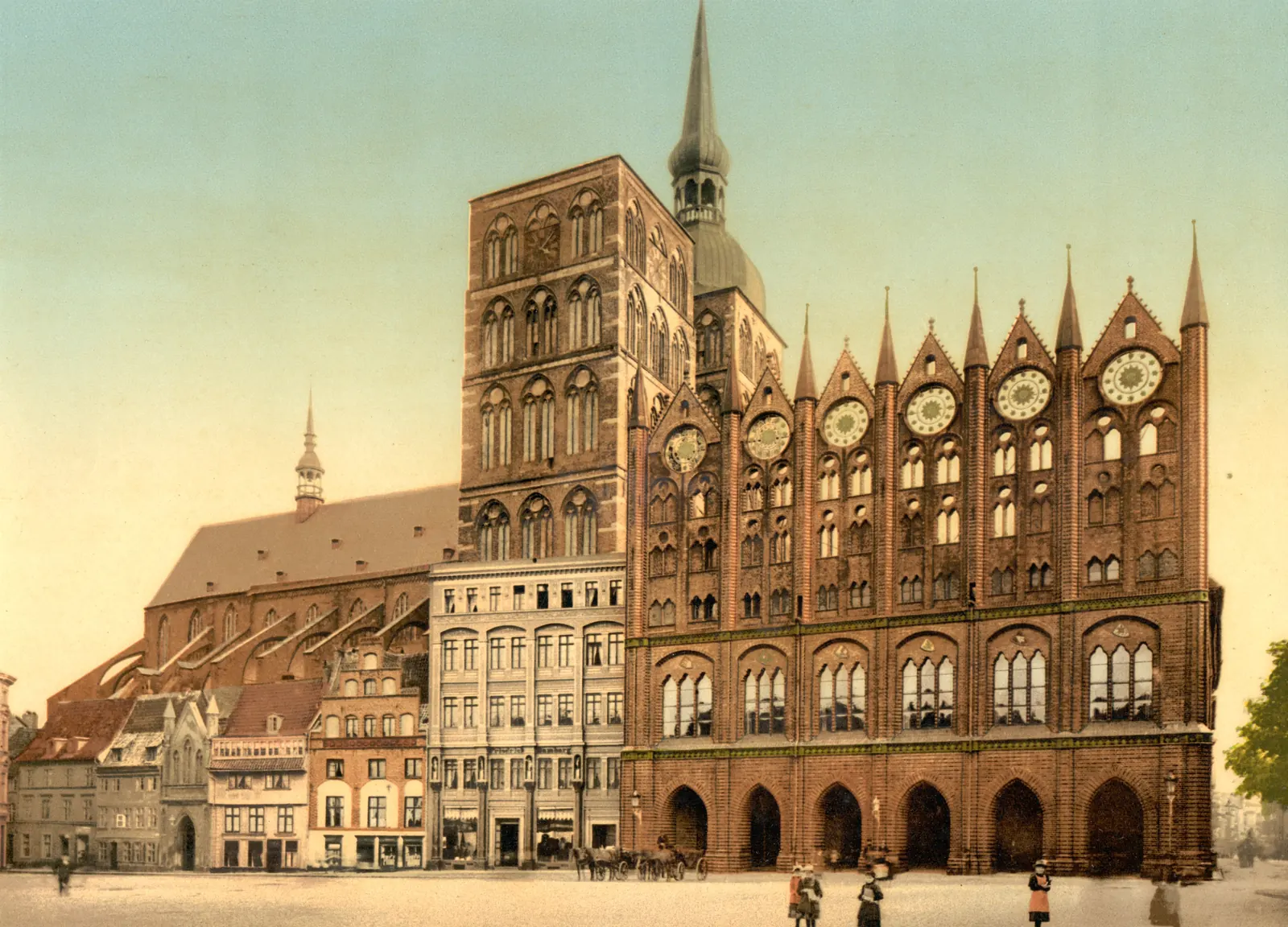 Colored photograph of a brick Gothic town hall with a screen of tall gables, pinnacles and round windows, a great church tower behind it