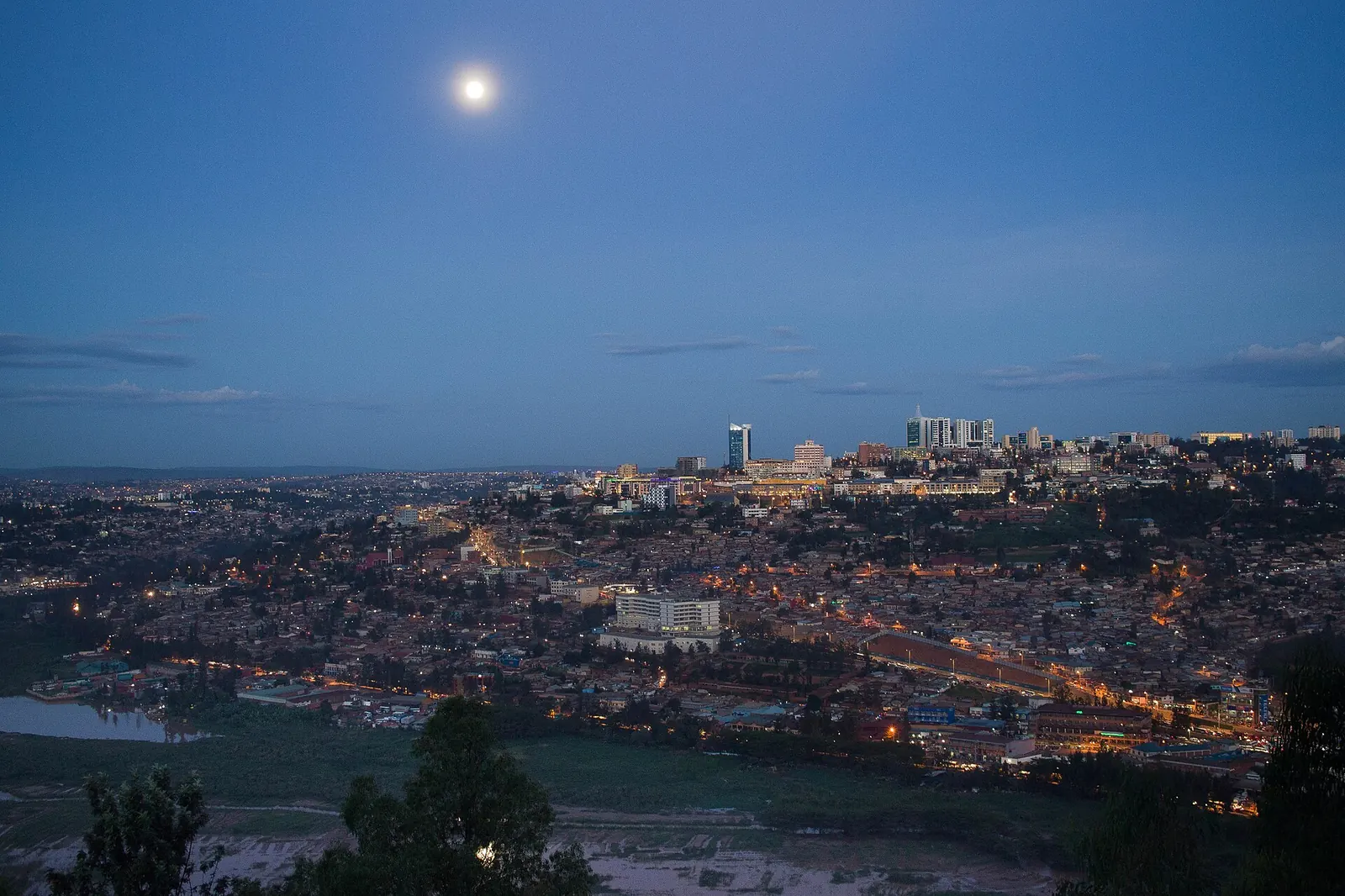 Kigali at dusk: lit towers downtown, hills of houses and a full moon above