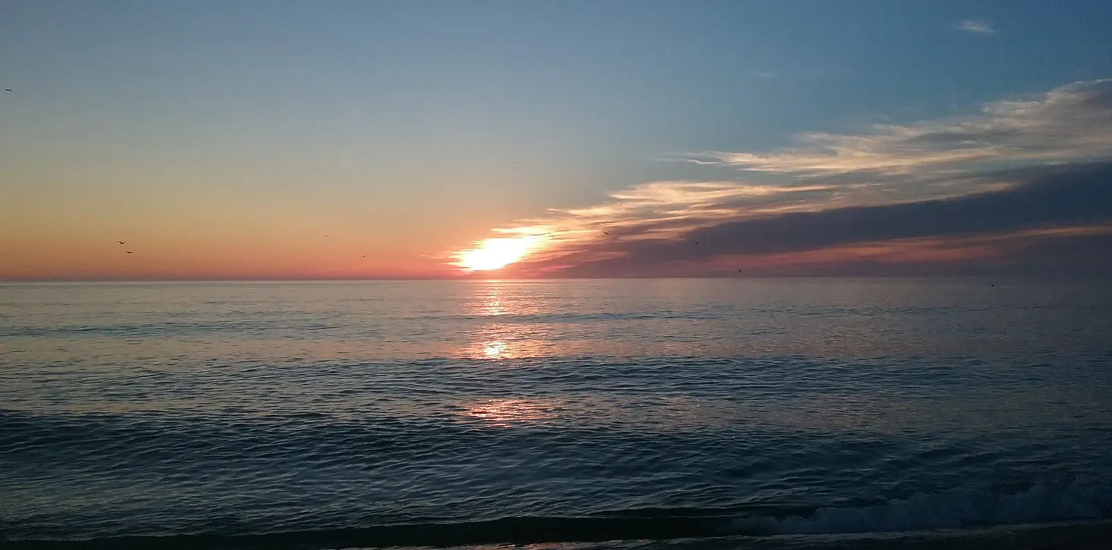 The sun rising out of the Atlantic beneath a long bank of cloud, its light running across calm water to the shore