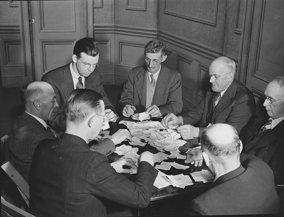 Seven men in suits seated around a table covered with folded paper slips, opening and sorting them
