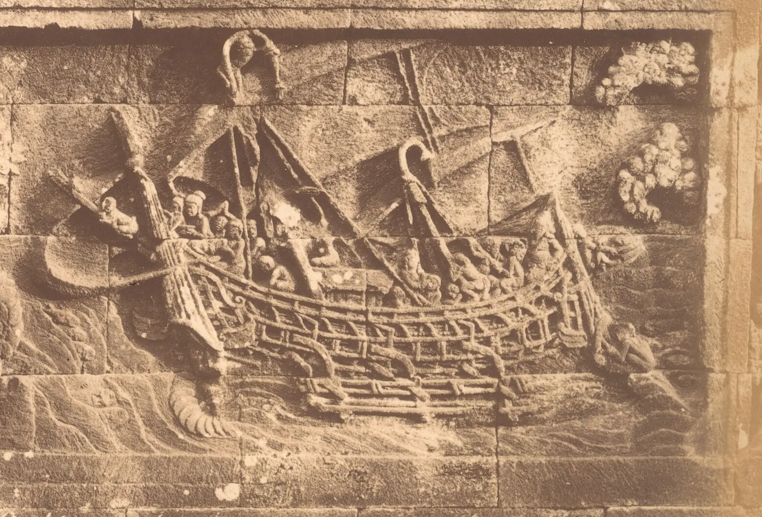 Stone relief of a large outrigger ship under sail, crowded with people, its masts, rigging and outrigger frame carved in detail
