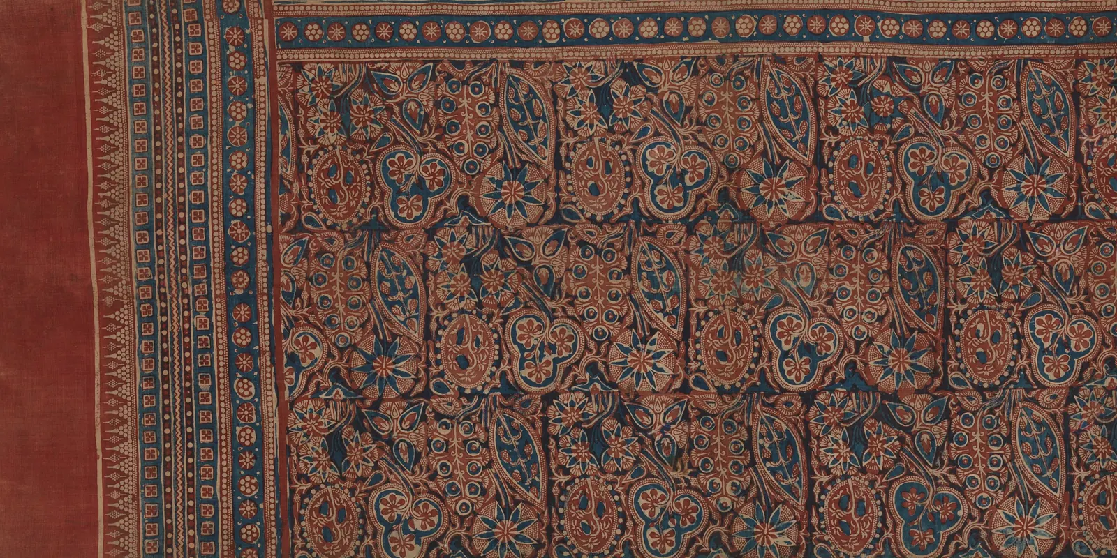 Long cotton cloth painted in red and indigo with rows of stylized trees, a plain red end panel edged with a band of small triangles, and patterned borders