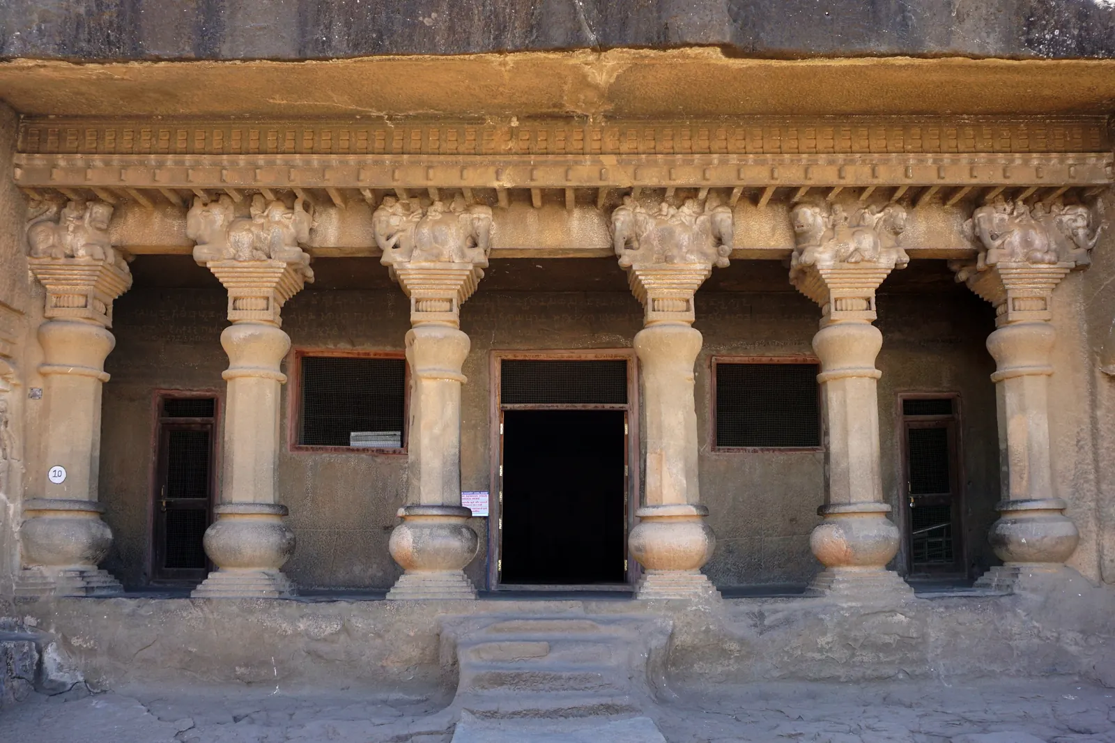 Rock-cut veranda with six pillars crowned by carved animals, three doorways and two windows in its back wall