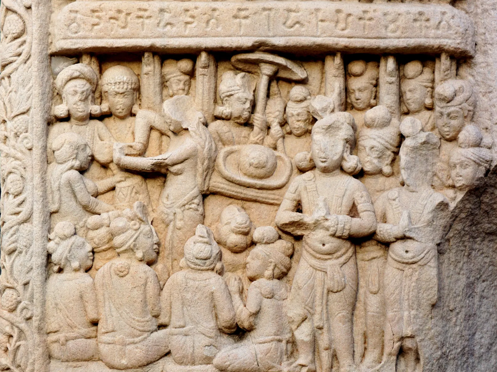 Sandstone relief crowded with standing and seated figures beneath a band of carved letters