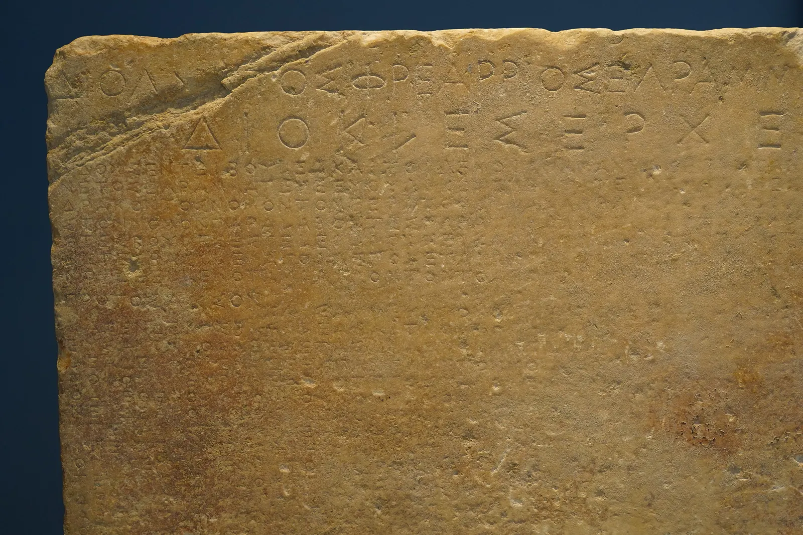 Top of a marble stele: two lines of large Greek letters above many rows of small letters set in an even grid