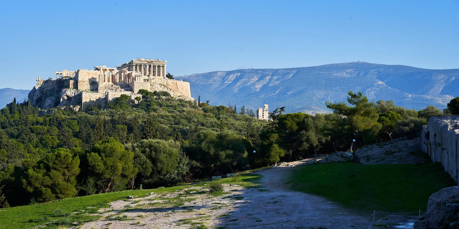 A rocky plateau in late sun with a cut stone wall at right, pines below and the Acropolis on its hill beyond